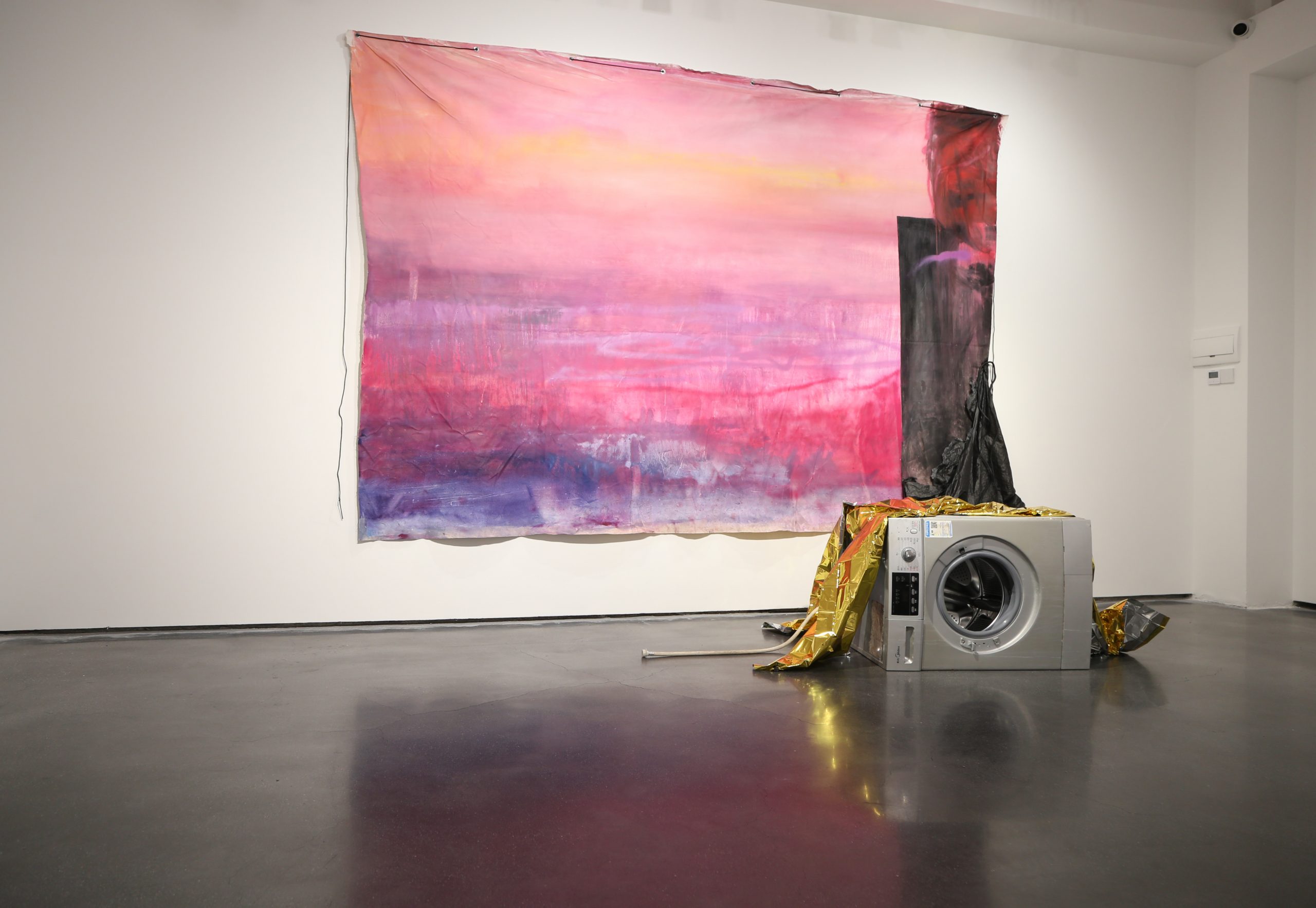 in the foreground a broken washing machine covered with a golden emergency blanket. in the background a large magenta abstract painting resembling a sunset